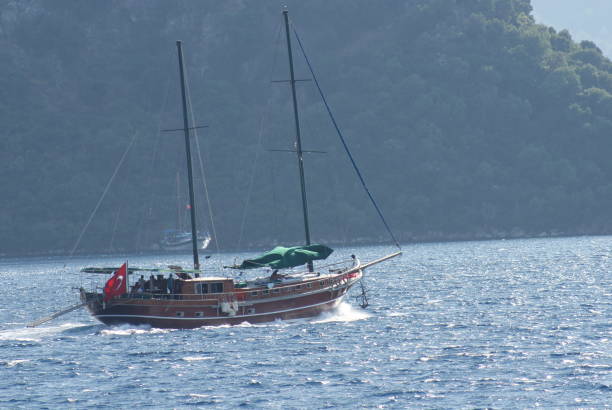 A beautiful sailboat glides over the sparkling blue waters near the coast of Marmaris, Turkey. The boat, with its white sails catching the wind, contrasts against the rugged, tree-covered mountains in the background. The scene captures the essence of a peaceful and adventurous sailing experience in the Mediterranean.