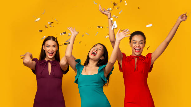istock Three Millennial Girls In Dresses Throwing Confetti Over Yellow Background 2177474872