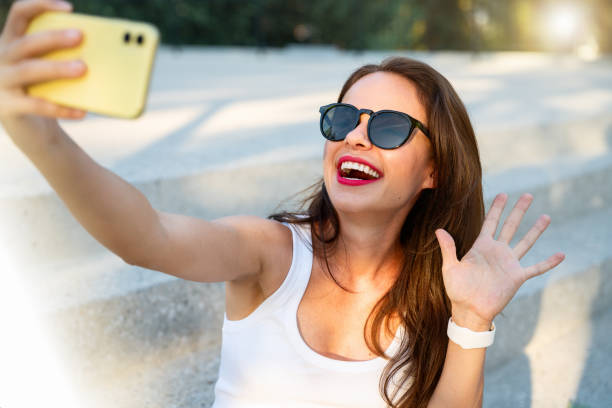 istock Woman in sunglasses sitting on steps outside, using her mobile phone to take a selfie or make an online video call. 2177481909