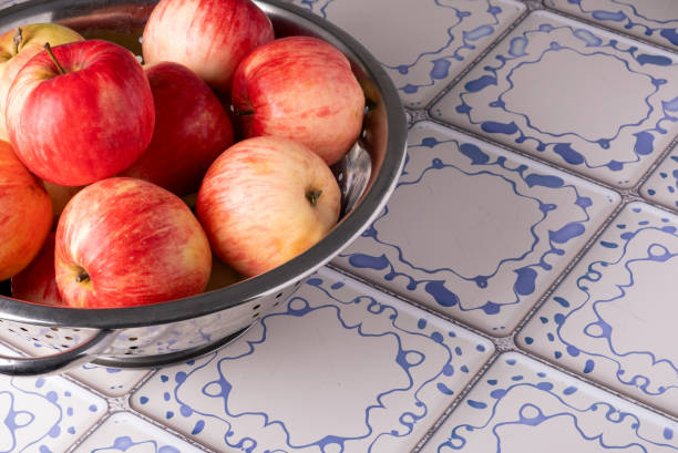 Ripe red apples lie in a metal colander on a table with a blue and white tile surface. Selective focus.