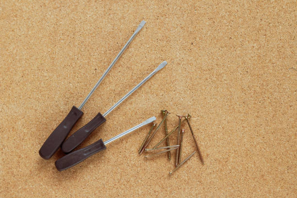 Three flathead screwdrivers with dark brown handles and a small pile of screws placed on a corkboard surface. The setup suggests a home repair or DIY project.