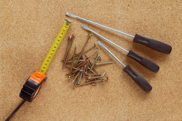 Three flathead screwdrivers with dark brown handles, measuring tape and a small pile of screws placed on a corkboard surface. The setup suggests a home repair or DIY project.