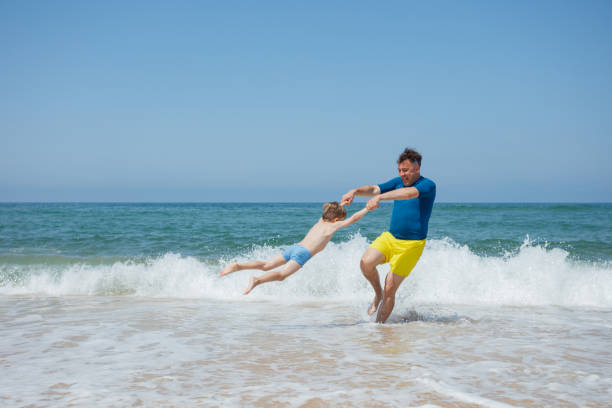A man in yellow shorts and a blue shirt is playing with a young boy on a beach, spinning him around, the ocean waves in the background