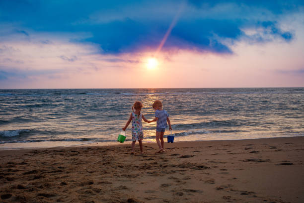 With the ocean waves gently lapping at their feet, two little kids walking together, hold hands in the amber glow of the sunset