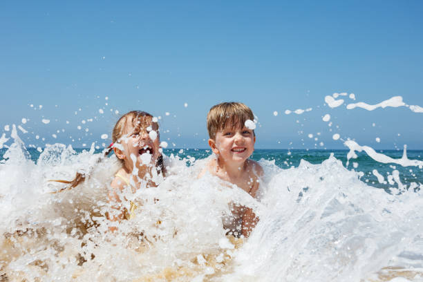 Two children laughing playfully as ocean waves splash around them on a bright, sunny day on summer vacations