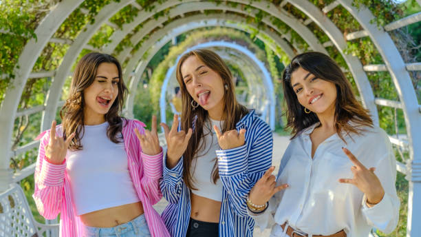 istock Three young female friends in a tree-filled park on a spring day, looking at the camera while making a metal hand sign 2177509765