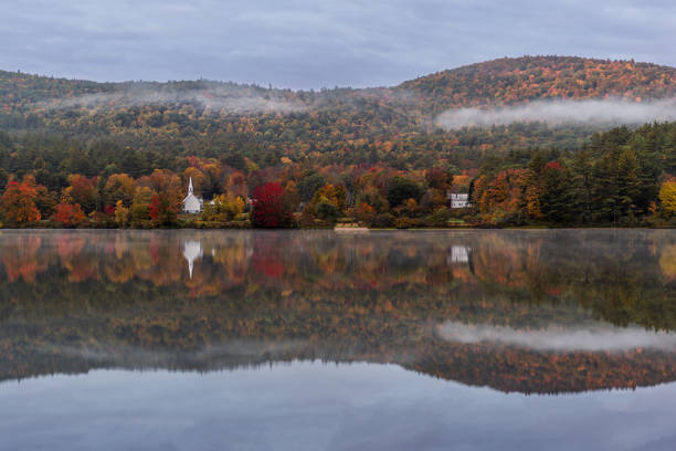 This photograph presents a peaceful lakeside scene, with the calm water reflecting the autumn-colored trees and a small white church in the distance. The misty sky adds a soft, tranquil atmosphere, and a thin layer of fog gently rests on the hillside behind the church. The vibrant fall foliage creates a beautiful contrast with the serene water and overcast sky, adding depth to the composition. The symmetry between the landscape and its reflection in the water enhances the calmness and natural beauty of the image.