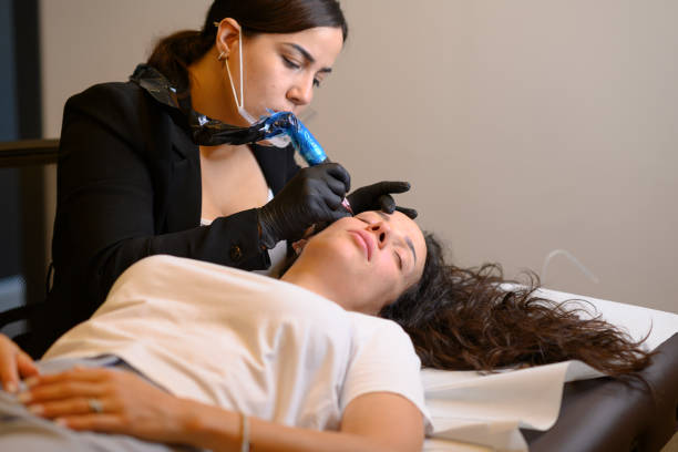 Beautician applying permanent eyebrow makeup with microblading machine on brunette hispanic 30 woman