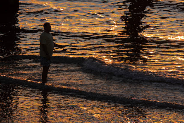 Port Aransas, TX - The early morning sun gives a warm, gold color to the Gulf of Mexico and the sand in a  high-angle photo of a male fishing under the Horace Caldwell pier at sunrise.