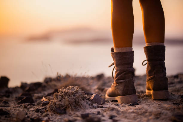 Close up of unrecognizable hiker in hiking boots standing on a hill above the sea at sunset. Copy space.