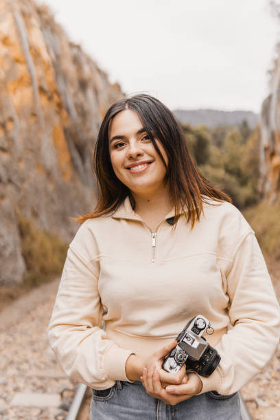 A young woman smiles while holding a vintage camera, with rocky canyon walls in the background