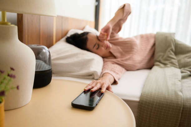 Sleeping asian woman turning off alarm on smartphone while being Waken up in the morning stock photo