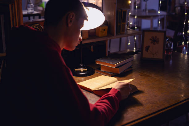istock Young man is reading an old book in the library under a table lamp 2177565656