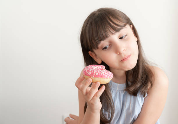 Little girl is eating pink doughnut with sprinkles in front of white background.