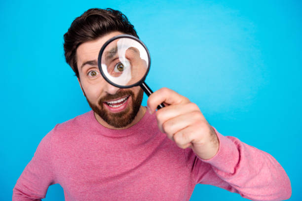 istock Joyful man with beard and pink sweater playfully looking through magnifying glass on bright blue background 2177572671