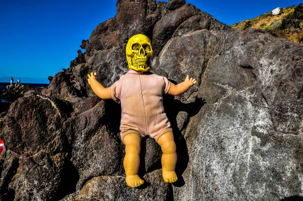 A doll with a yellow skull on its head is sitting on a rock. The doll is wearing a white outfit and he is looking up at the sky. The scene has a creepy and eerie mood, as the doll's skull