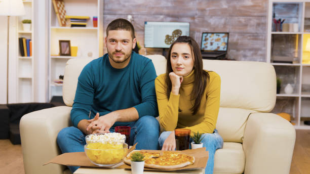 istock Couple sitting on couch laughing while watching tv and eating pizza 2177581860