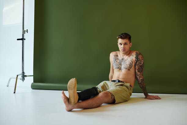 A young man sits casually in a studio, showcasing his tattoos and artificial limb with pride.