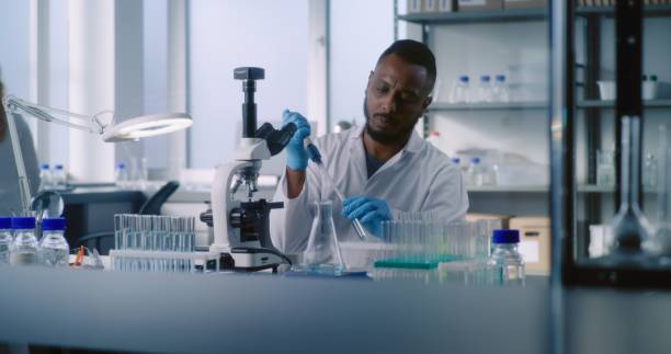 istock African American microbiologist takes liquid from test tube, looks under microscope 2177597801