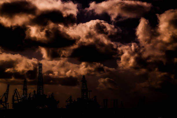 The sky is dark and cloudy, with a few oil rigs visible in the background. The oil rigs are silhouetted against the sky, creating a sense of depth and perspective. Scene is somber and somewhat ominous