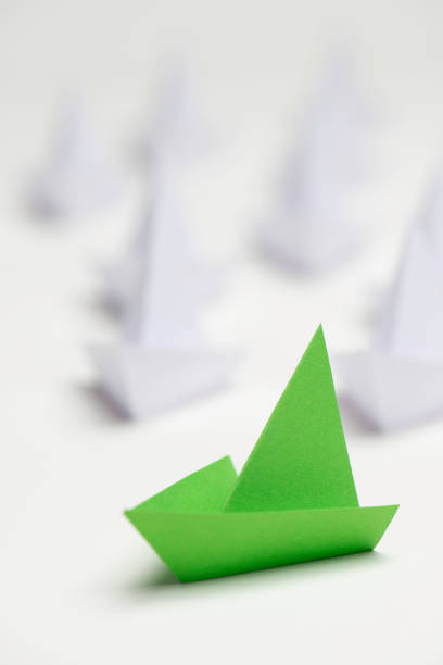 A conceptual image of paper boats, with a pink boat standing out among white ones, symbolizing leadership, individuality, and standing out from the crowd. The green boat represents innovation and forward-thinking leadership, while the uniform white boats reflect conformity. Ideal for illustrating themes of leadership, creativity, innovation, and differentiation.