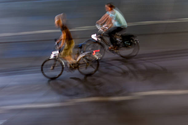 Panning shot capturing two cyclists riding through city streets at night. The blurred motion creates a dynamic and energetic urban scene.