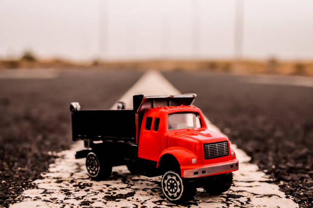 A red toy dump truck is sitting on a road. The truck is small and has a black cab. The road is empty and the sky is cloudy