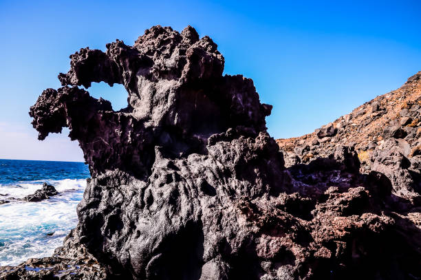A large rock with a hole in the middle of it. The rock is surrounded by water and the sky is blue