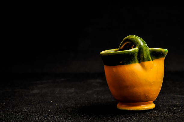 A small green and yellow bowl sits on a black table. The bowl is made of clay and has a green handle. The bowl is empty and he is a decorative piece