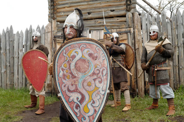 Four Viking reenactors holding shields and weapons, donning traditional armor and mail, standing in front of wooden fortress, exuding readiness for combat
