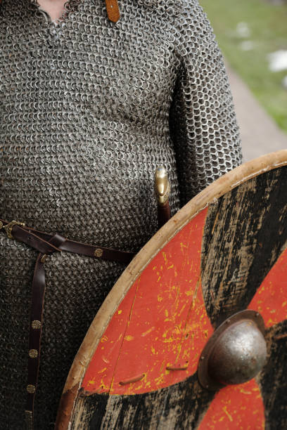 Close-up of person wearing chainmail armor and carrying round wooden shield with metal boss and painted design on green grass background