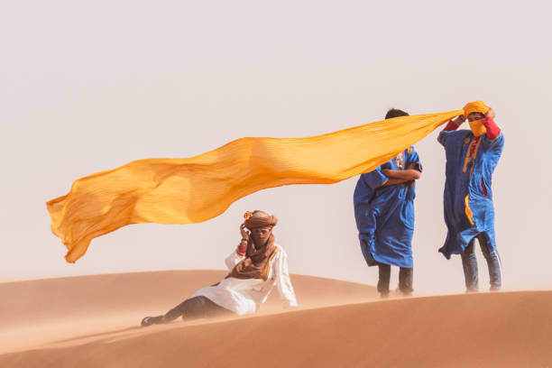 Moroccan wearing a turban at the top of a dune, accompanied by two friends. There is a strong sandstorm, which makes the scarf he is putting on show all its beauty by moving in the wind.