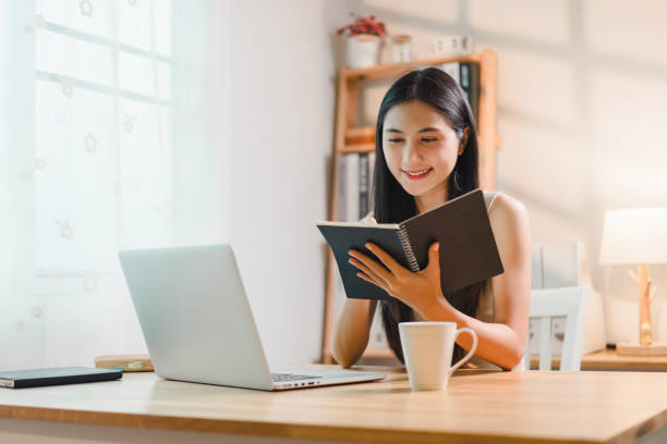 istock Young asian woman studies at her home workspace engaging with her notebook while a laptop and coffee cup sit nearby, creating a warm and inviting atmosphere. 2177685353