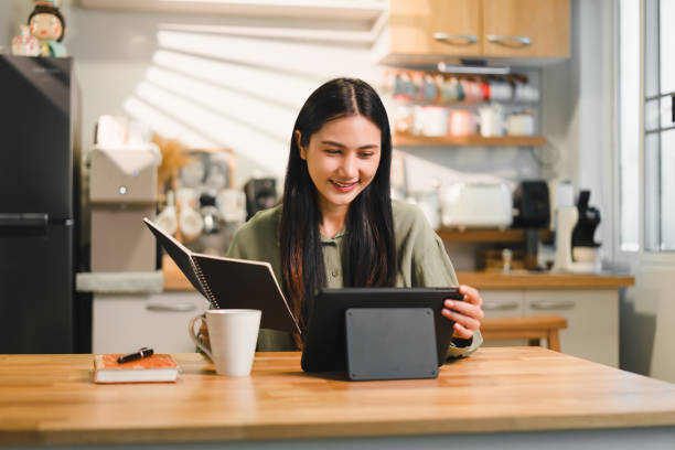 istock Young woman sits at wooden table in cozy kitchen smiling while engaging with her tablet and taking notes in a notebook enjoying her coffee. 2177685395