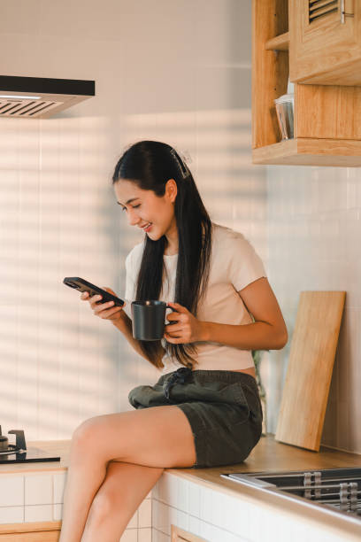 istock Smiling woman sitting on kitchen counter holding coffee mug and using smartphone enjoying a relaxed morning in a cozy kitchen. 2177685540