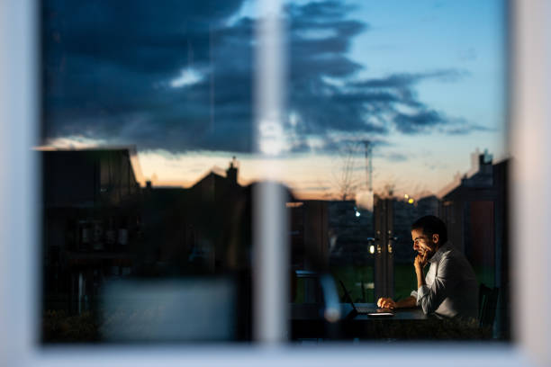 Businessman working from home late at night using laptop computer Tired businessman works late at his home office, lit by the sunset outside. Stress is visible as he types on his laptop, balancing work and life office window night stock pictures, royalty-free photos & images