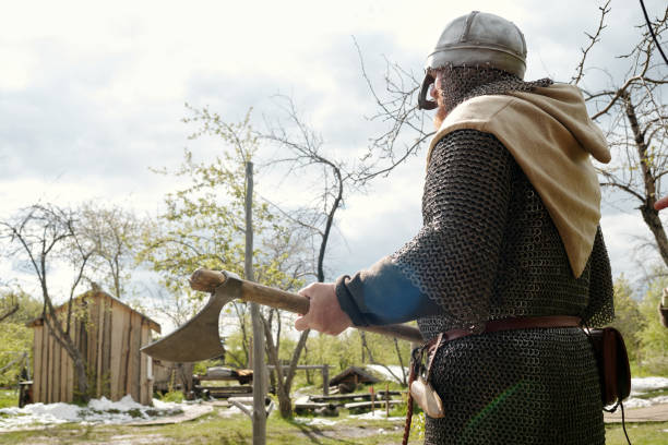 Medieval knight wearing chainmail armor and helmet holding an axe and standing in a rustic village on a sunny day