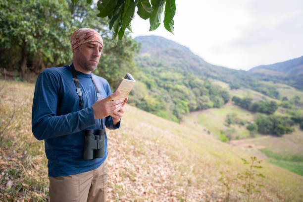 Man observing birds looking through binoculars and using field guide book for bird identification. Listening to birdsong in nature.Adventure travel activity and outdoor trekking lifestyle.