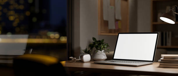 istock A laptop mockup on a table by the window in a room at night, illuminated by a table lamp. 2177695872