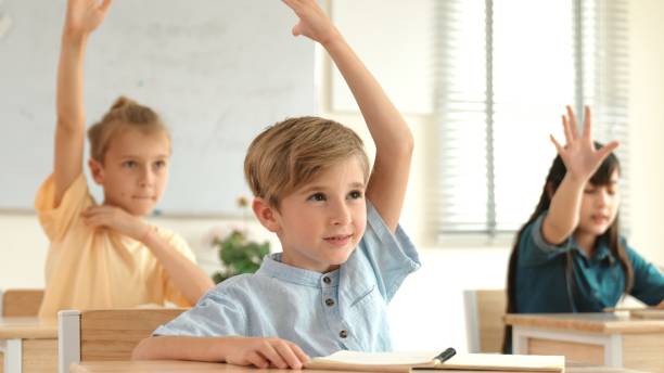 Smart caucasian boy raising hand for asking teacher at classroom. Pedagogy.