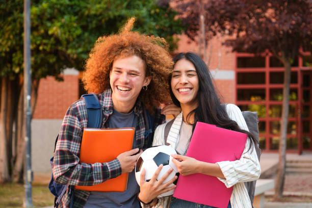A multiracial group of high school or college students in a library reading room, carrying book bags and textbooks, smiling at the camera.  The young man standing in the middle, wearing eyeglasses and carrying a book and bookbag looks like he is the leader of the group.