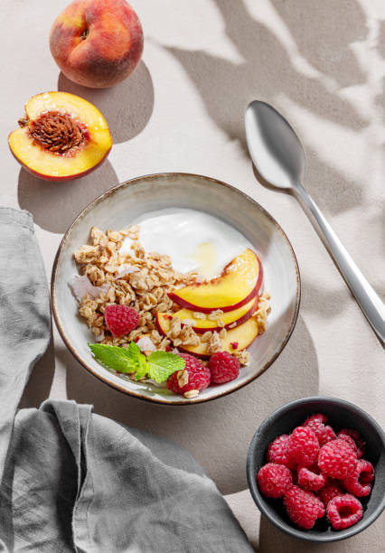 istock Greek yogurt with oat granola or muesli, peach and raspberry in a bowl on a light background with fresh fruits, berries and morning shadow. 2177738245