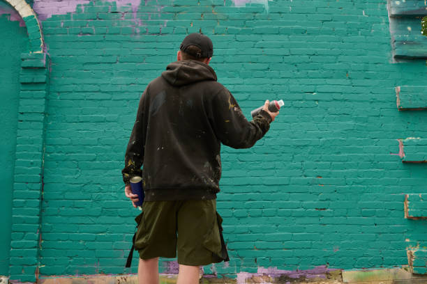 Back view of young man in black hoodie standing in front of green brick wall of building before spraying paint and creating artowrk