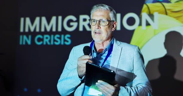 istock Seminar, speaker and man at screen for global immigration presentation with microphone, tablet and information. Ideas, change and mature presenter at podium for conference on world refugee crisis 2177825907