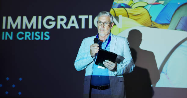 istock Seminar, speaker and man on stage for global immigration presentation with microphone, tablet and information. Ideas, change and mature presenter at screen for international refugee crisis conference 2177825921