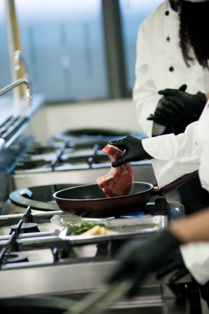 Professional multiracial student chef wearing black glove, white chef's hat and cloth, preparing the fresh beef steaks in the cooking pan seasoned with dill and butter during learning cooking class in modern restaurant. Cooking school
