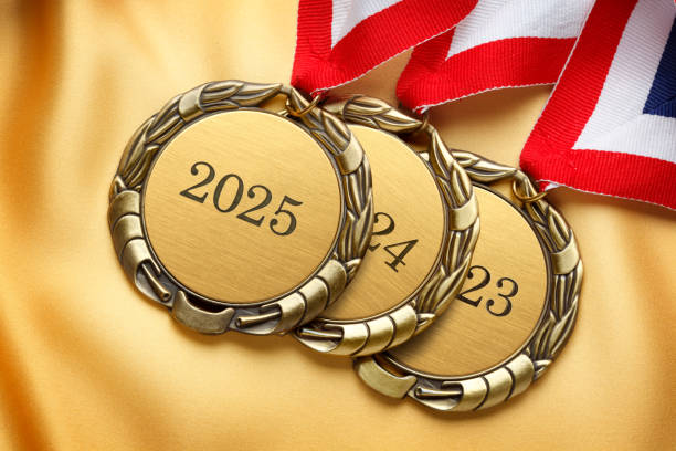A stack of three gold medals resting on gold satin fabric.  The top medal is engraved with 2025.  A red, white, and blue ribbon is attached to each medal.