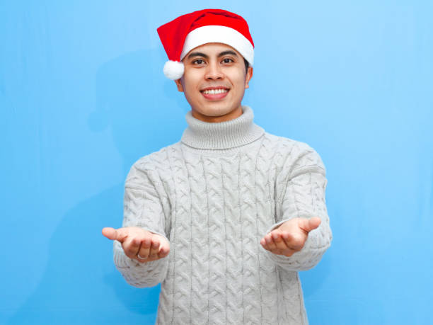 istock Smiling young Asian man wearing a sweater and Santa hat, showing something with empty hands, isolated on a blue background. Perfect for holiday promotions and festive advertisements. 2177871067