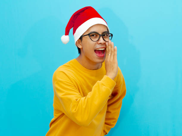 istock Asian man wearing a Christmas hat, whispering something with a happy expression. Perfect for festive advertising, holiday promotions, and cheerful seasonal content. 2177881541