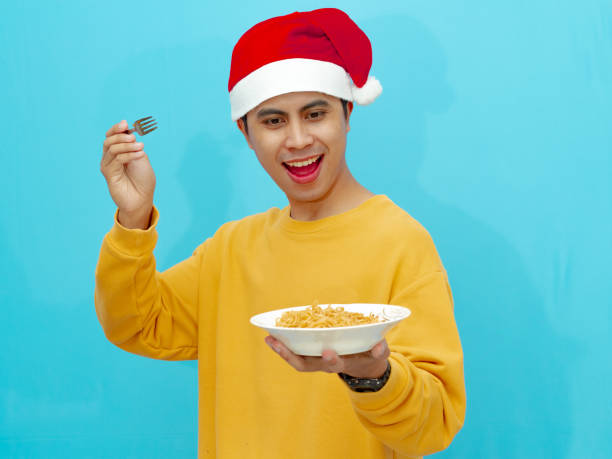 istock A happy Asian man in a yellow sweater and Christmas hat smiling while holding a fork, looking at a plate of fried noodles. Isolated on a blue background. 2177881904